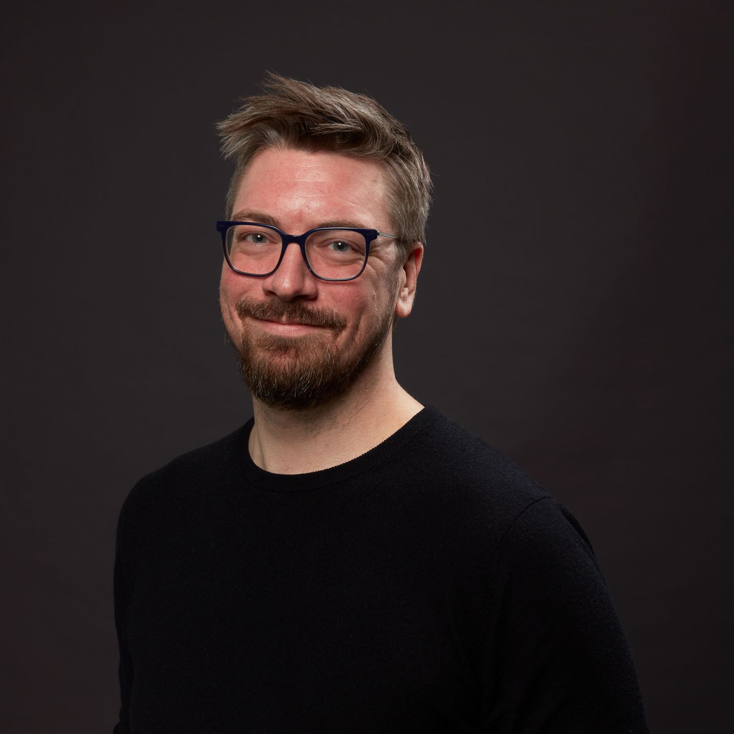 Headshot of Dylan Garret, smiling and facing forward, wearing black sweater and glasses on solid dark gray background