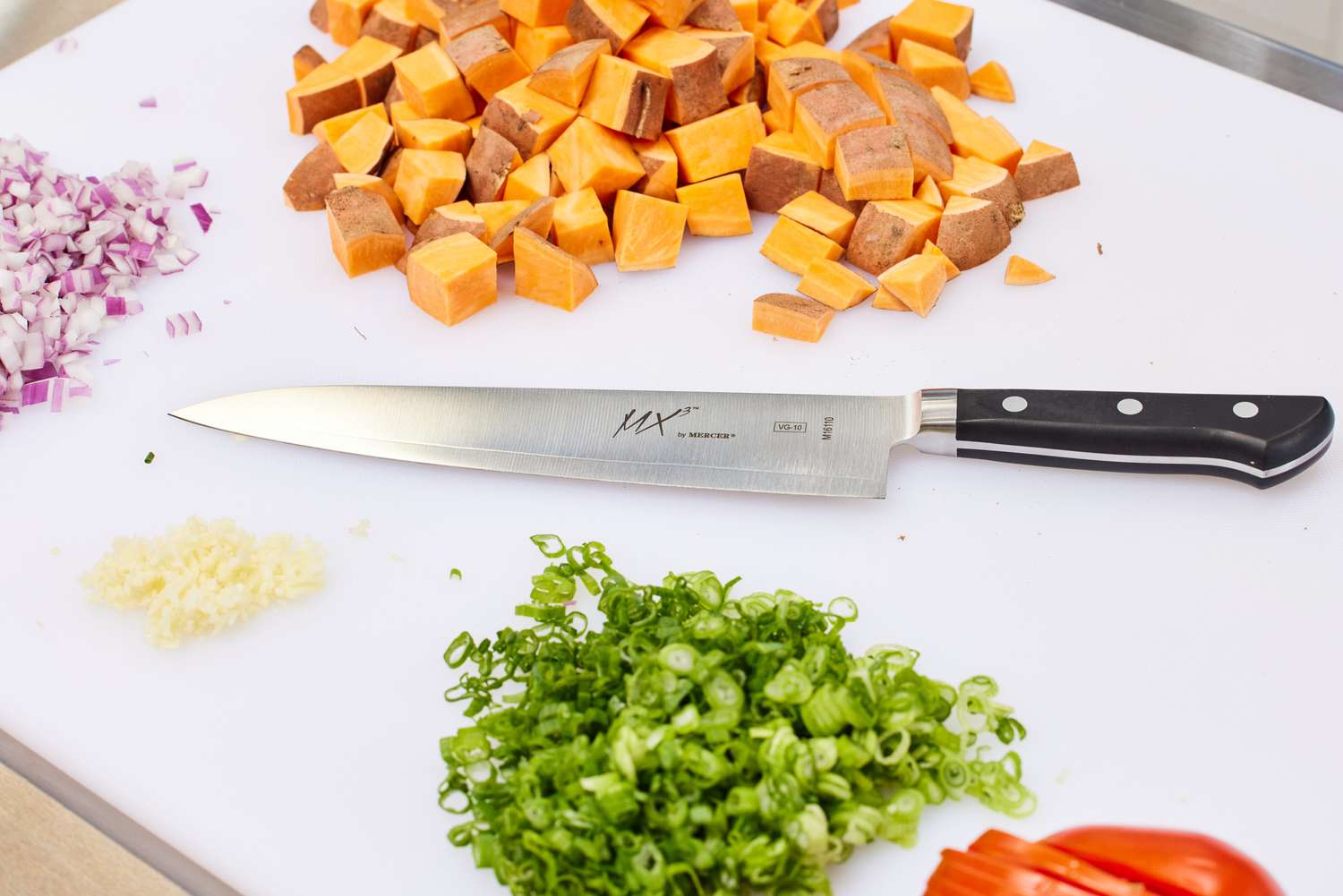 A Mercer MX3 8.25-Inch Chef Knife lying on a white cutting board next to cut vegetables