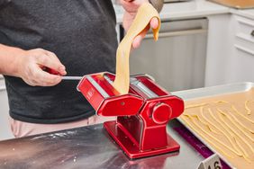 Person feeding pasta dough through red Marcato Atlas 150 Pasta Machine next to tray of cut pasta strands