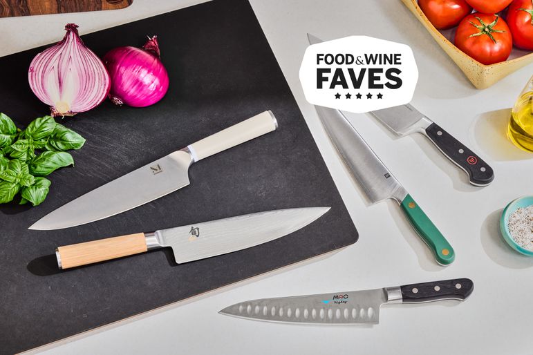 Several chef's knives on a counter with a cutting board, tomato, onion, and basil