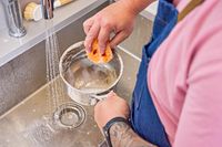 A person using a sink and sponge to clean a pot from the Le Creuset Tri-Ply 10-Piece Stainless Steel Cookware Set