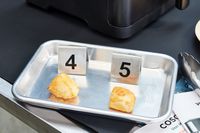 Two chunks of cooked chicken displayed on a stainless steel tray with a numbered label behind each one