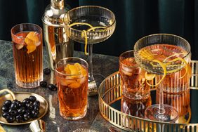 Various cocktails, a cocktail shaker, and black olives on a golden tray and marble surface. 