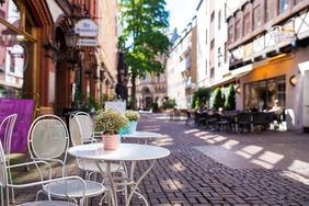 White chairs and tables with flower pots on a cobblestone street lined with restaurants and storefronts