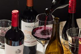 Red wine being poured into a glass while surrounded by bottles and glasses of wine. 