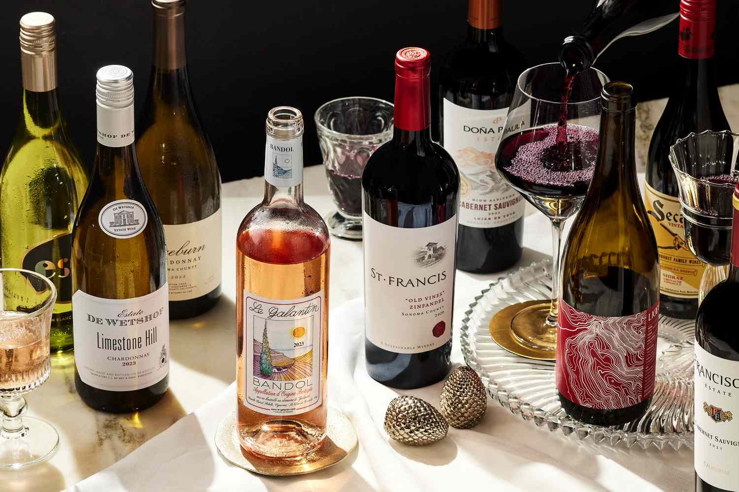 Various bottles of red, white and rosé wines on a table top while a glass of red wine is being poured. 