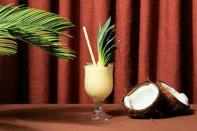 Pina colada cocktail in a glass standing under a palm tree branch, next open to a cracked open coconut and in front of a brownish-red curtain. 