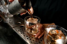 bartender pours negroni cocktail into glass with large piece of ice on the bar counter.