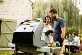 Two people standing in front of a Weber Spirit II E-310 while two other people are sitting at a table in the background