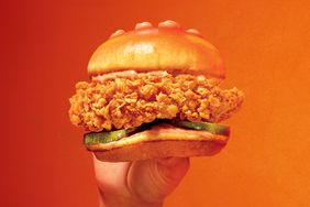 A hand holding a Popeyes Ghost Pepper Chicken Sandwich in front of an orange background. 