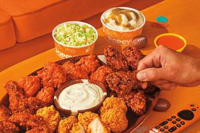 A person picking up a chicken wing from Popeye's next to a remote control, mini footballs, coleslaw and mashed potatoes.