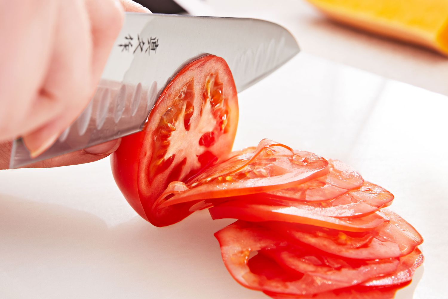 Person using a chef's knife to slice a tomato on a white cutting board