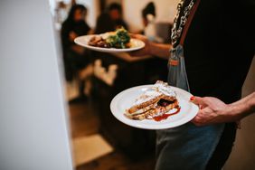 Beautifully presented food is plated up in a commercial kitchen, ready to be served to customers. A waiter carries the plates from the kitchen into the cafe.