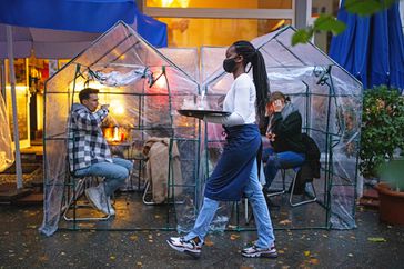 People sitting inside plastic tents at outdoors cafe with restaurant server wearing face mask carrying a serving tray.