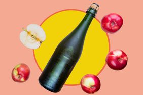 A bottle of hard apple cider and apples on a yellow and peach colored background. 